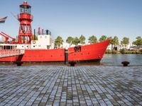 the large boat is sitting next to the light house and harbor tugboats on brick path near the river