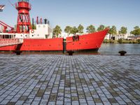 the large boat is sitting next to the light house and harbor tugboats on brick path near the river