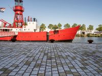 the large boat is sitting next to the light house and harbor tugboats on brick path near the river