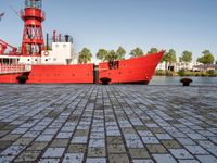 the large boat is sitting next to the light house and harbor tugboats on brick path near the river