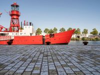 the large boat is sitting next to the light house and harbor tugboats on brick path near the river