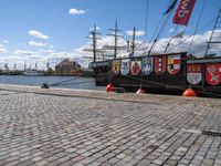 a ship tied to a dock on a brick walkway near water and flags and red buoys