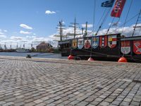 a ship tied to a dock on a brick walkway near water and flags and red buoys