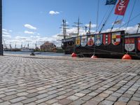 a ship tied to a dock on a brick walkway near water and flags and red buoys