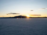 the sun is setting over a field covered in snow and mountains are visible behind it