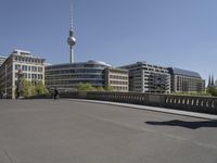 a boy skateboards across the bridge at berlin's tv tower, and some people are seen in the distance