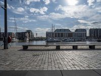 a bench on a paved street overlooking the water with a sun beam and other buildings in the distance