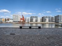 a park bench sitting on the edge of a dock with buildings behind it and blue skies