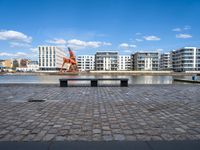 a park bench sitting on the edge of a dock with buildings behind it and blue skies