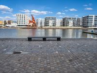 a park bench sitting on the edge of a dock with buildings behind it and blue skies