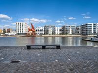 a park bench sitting on the edge of a dock with buildings behind it and blue skies