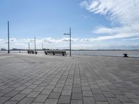 a long park bench sitting on the side of a brick road by water under a cloudy blue sky