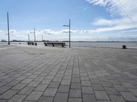 a long park bench sitting on the side of a brick road by water under a cloudy blue sky