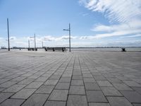 a long park bench sitting on the side of a brick road by water under a cloudy blue sky