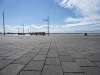 a long park bench sitting on the side of a brick road by water under a cloudy blue sky