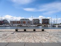 a stone bench on the ground near a body of water with buildings in the background