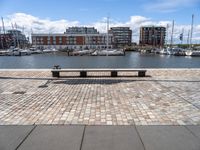 a stone bench on the ground near a body of water with buildings in the background