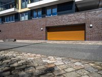 a sidewalk near some cars and a brick building with a orange garage door on the side of it