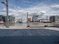 a park bench in front of some buildings and water with boats near them, in a park next to an industrial site