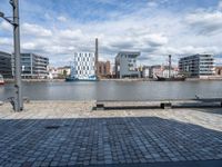 a park bench in front of some buildings and water with boats near them, in a park next to an industrial site