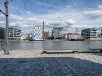 a park bench in front of some buildings and water with boats near them, in a park next to an industrial site