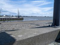 an empty concrete bench near some water and buildings in the background of a stone walkway