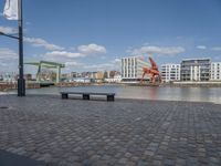 benches sitting on a brick floor by the water in the city with tall buildings and cranes