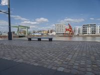 benches sitting on a brick floor by the water in the city with tall buildings and cranes