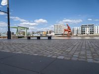 benches sitting on a brick floor by the water in the city with tall buildings and cranes