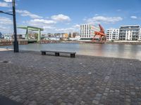 benches sitting on a brick floor by the water in the city with tall buildings and cranes