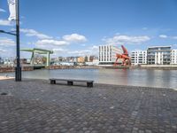 benches sitting on a brick floor by the water in the city with tall buildings and cranes