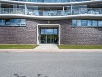 a large building is next to the street that leads up to it, and shows an archway leading through to a staircase, with a long white railing and curved door and grass field on either side walkway