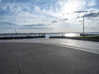 a skateboarder jumps on the pavement in front of a pier in sunny weather