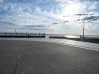 a skateboarder jumps on the pavement in front of a pier in sunny weather