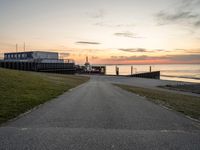 a long, paved road is near a grassy shoreline next to a pier in the evening