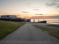 a long, paved road is near a grassy shoreline next to a pier in the evening