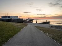 a long, paved road is near a grassy shoreline next to a pier in the evening