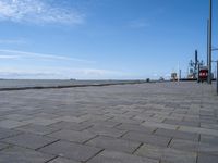 the sidewalk has brick pavers on it near a water front and the lighthouse in the distance