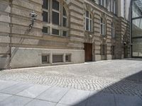 a brick sidewalk in front of a large building with windows on it and the ground is cobblestone