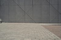 a brick walkway in front of an unfinished building with a bench outside it and in the foreground is a square pattern with squares of gray tiles on it