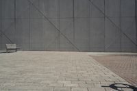 a brick walkway in front of an unfinished building with a bench outside it and in the foreground is a square pattern with squares of gray tiles on it