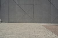 a brick walkway in front of an unfinished building with a bench outside it and in the foreground is a square pattern with squares of gray tiles on it