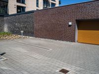 two men with orange safety helmets riding skateboards in front of a building, a yellow garage door and a brick sidewalk