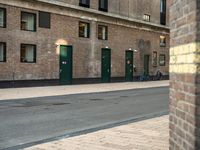 a couple of men walking past a brick building with several green doors on the other side