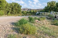 a bridge that crosses over some dirt road by the woods and grass of some bushes and flowers