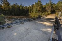 a bridge across a small river next to trees and dirt ground with benches in front