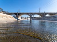 a large bridge spans over a shallow river between two buildings with power lines above it