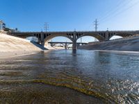 a large bridge spans over a shallow river between two buildings with power lines above it