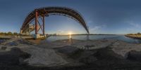 a bridge spans over the water near a shoreline at sunset time with a bright reflection of the sun coming out behind