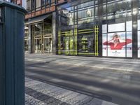 the large outdoor store front on the street corner is painted a bright yellow stripe and features advertises on glass and a poster